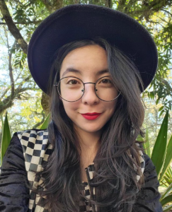 Foto da Niko olhando diretamente para a câmera, com óculos, chapéu e batom vermelho. Leve sorriso nos lábios.